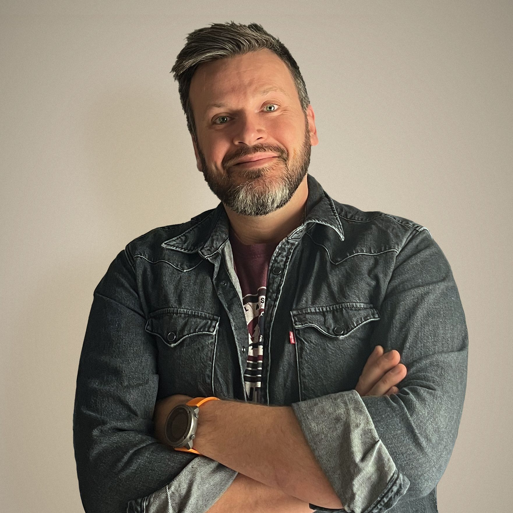 Portrait of the artist, Michael McAleer, standing with arms crossed in front of a sepia background. He's wearing a denim jacket and has short greying hair and a trim beard.